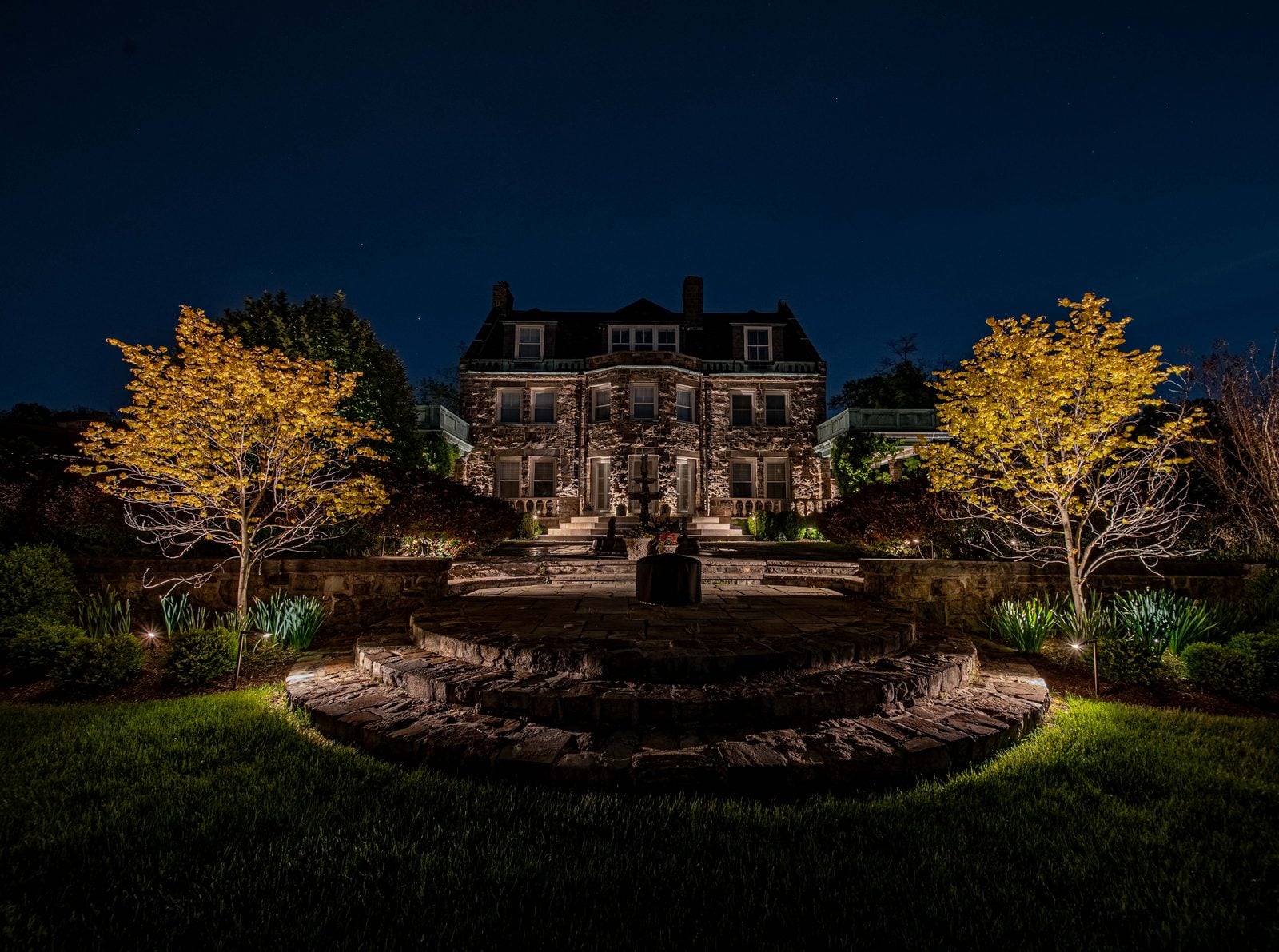 A stone manor at night, its facade washed with warm light and two specimen trees uplit either side of a circular fountain court